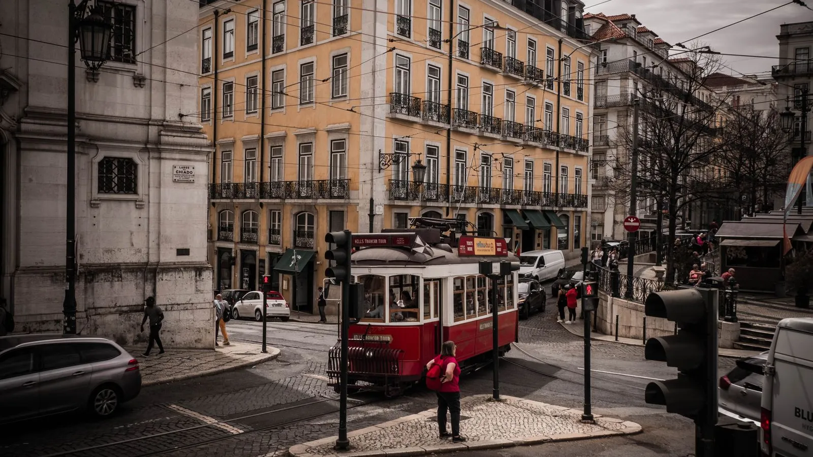 Elétrico vermelho a atravessar o Largo do Chiado, em Lisboa, com peões nos passeios e prédios pombalinos ao fundo.