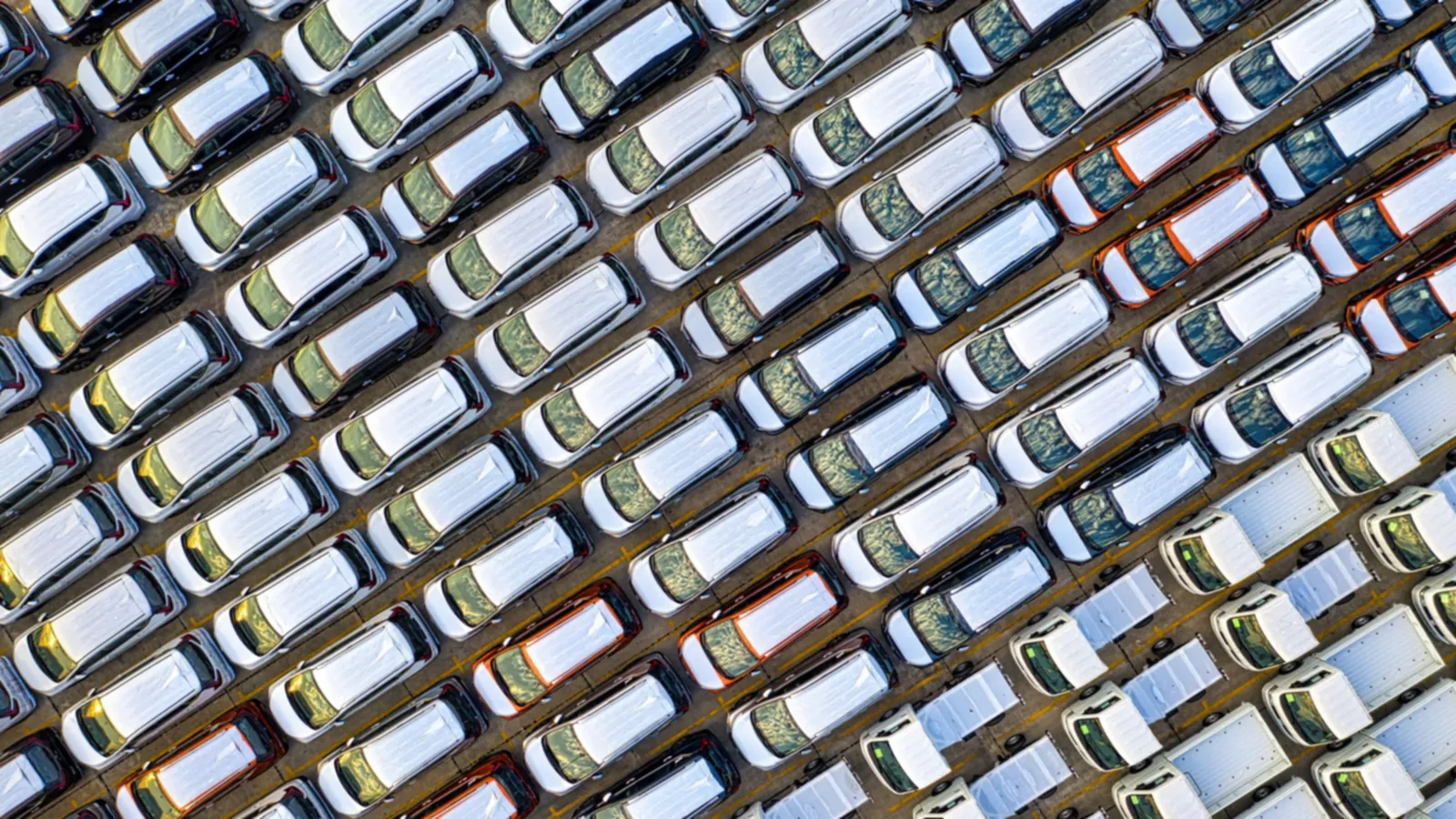 Vista aérea de dezenas de carros novos estacionados em filas diagonais, na maioria brancos, com algumas carrinhas de caixa aberta num dos cantos