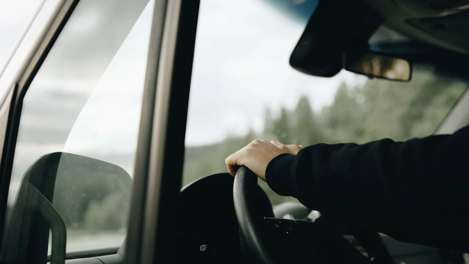 Mão de um motorista ao volante de um carro, vista do interior, com a estrada e árvores desfocadas ao fundo